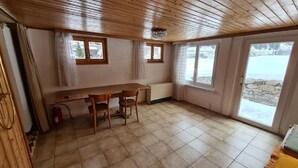 Een kamer met een houten plafond, betegelde vloer, een tafel met stoelen en grote ramen voorzien van gordijnen.