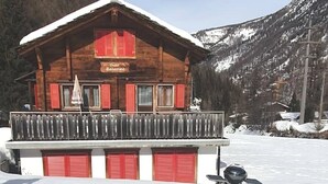 Een houten chalet met balkon en rode luiken, omgeven door besneeuwde bergen.