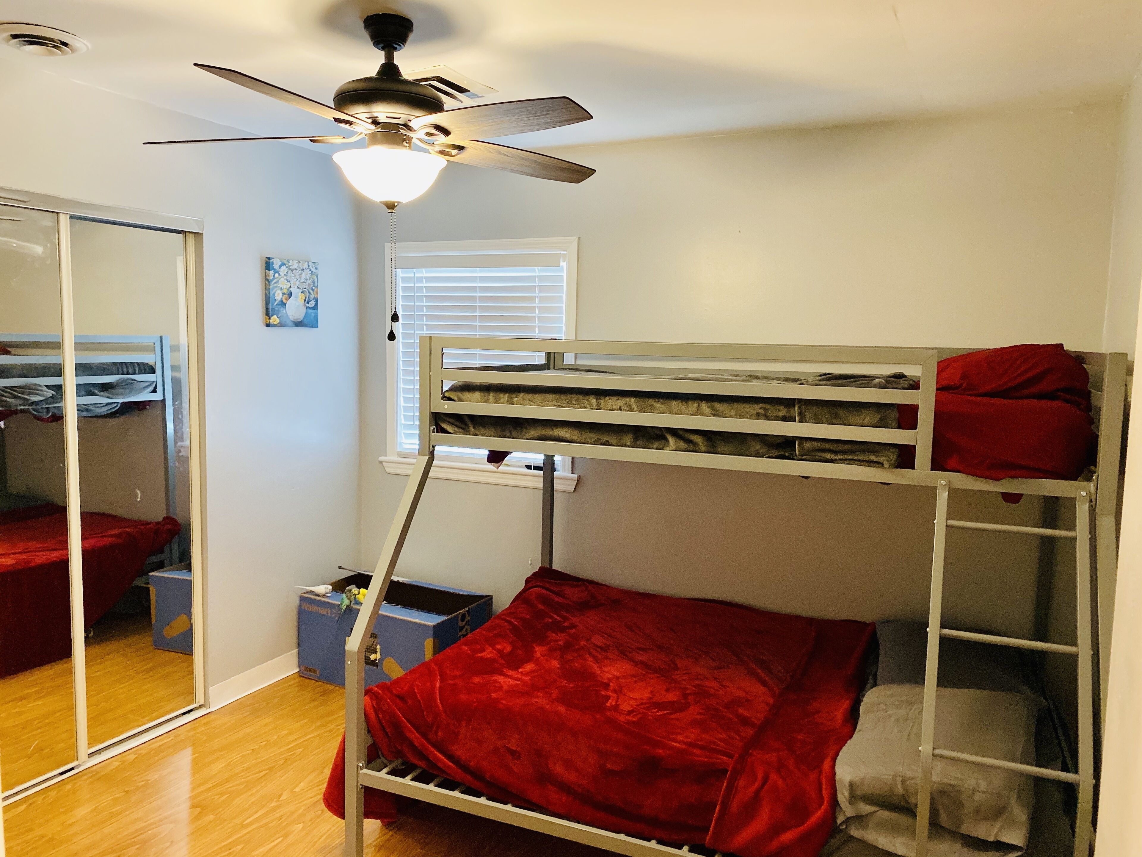 A bunk bed room with a ceiling fan, a window with blinds, and a mirror on the left wall.