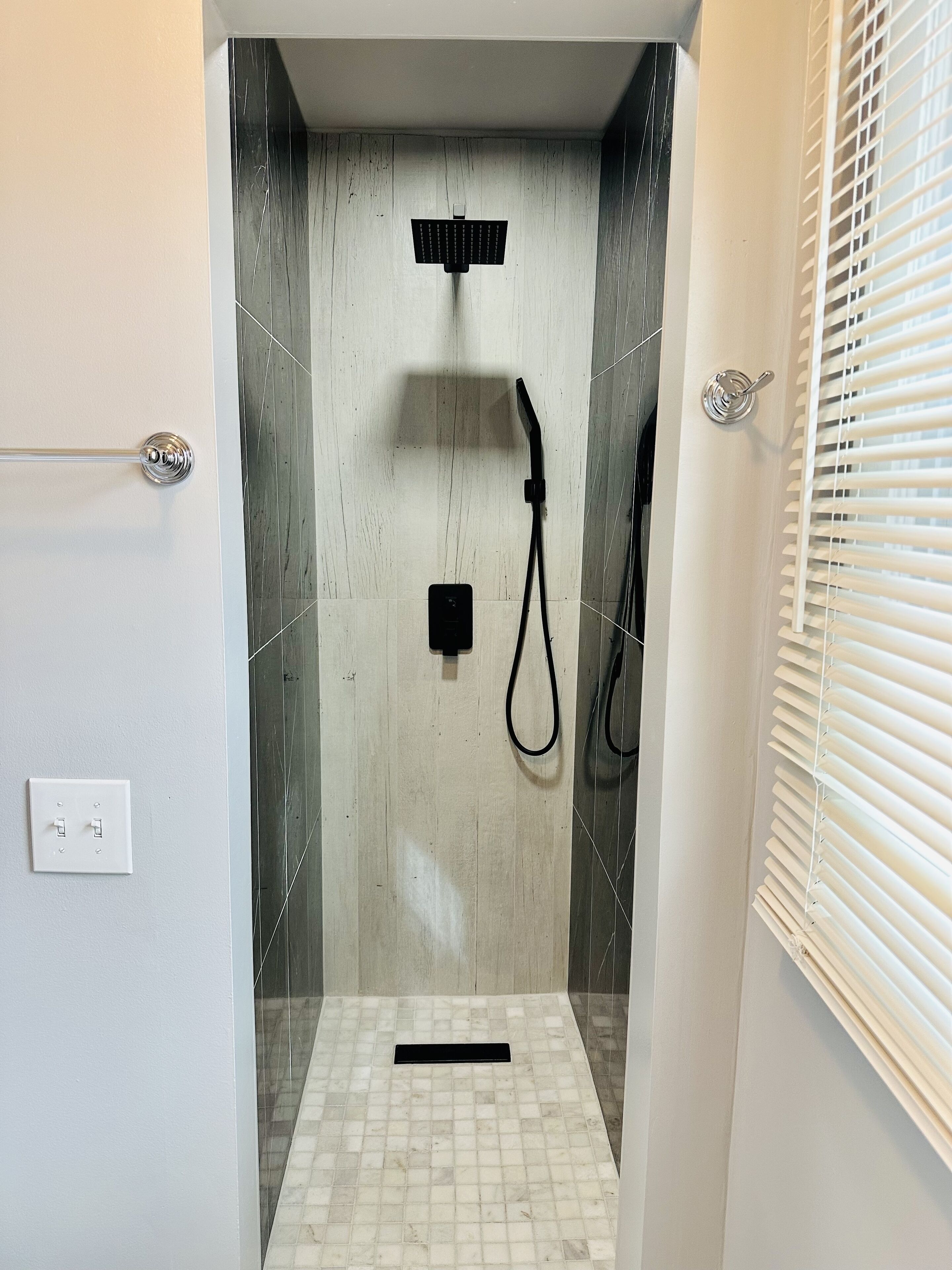 A shower area with a tiled floor, a handheld showerhead, and a wall-mounted control panel.