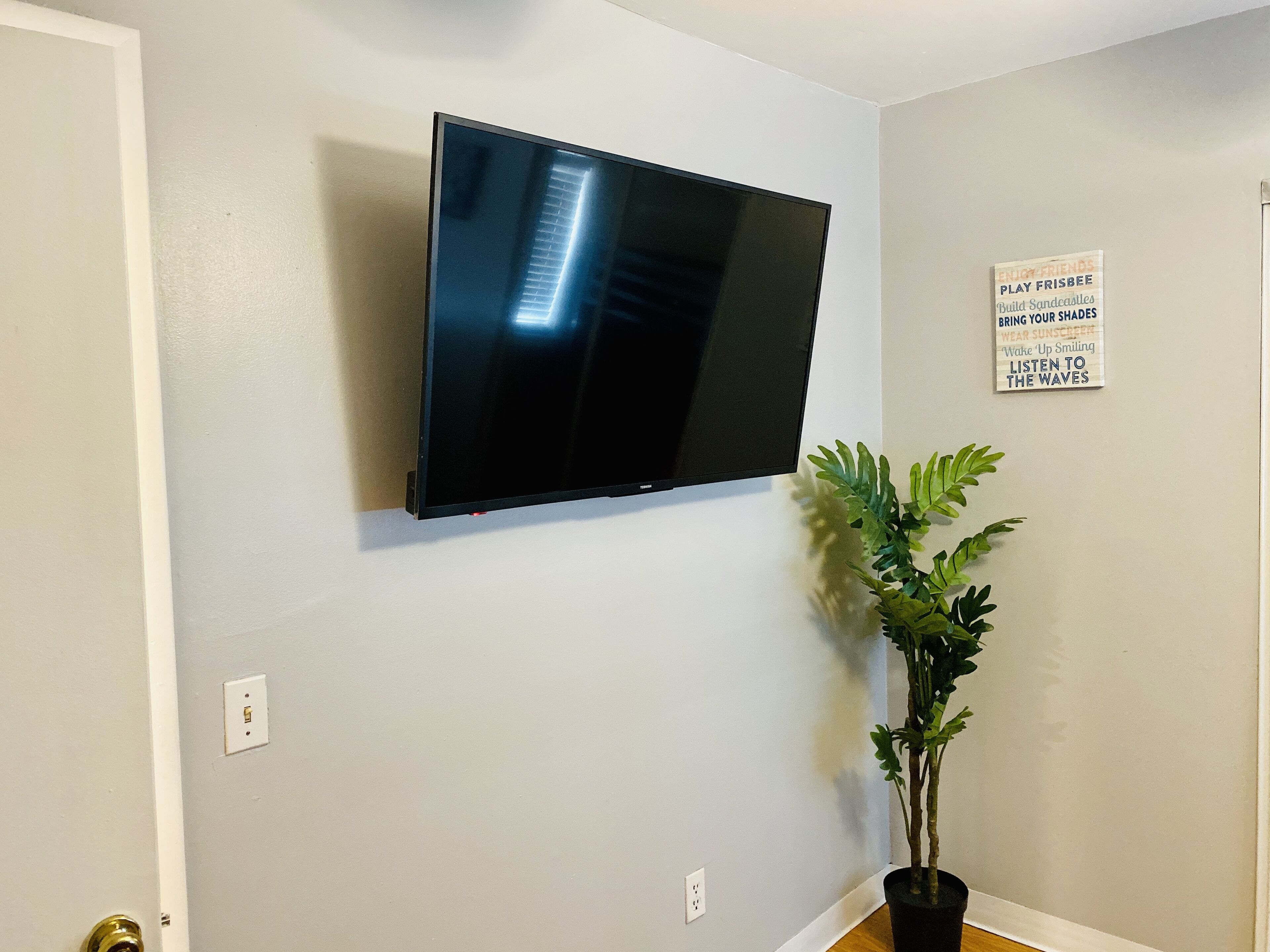 A wall-mounted flat-screen TV, a potted plant, and a motivational poster on a plain wall.