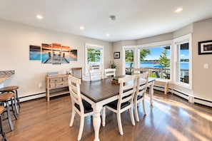 A dining area with a wooden table and chairs, a kitchen area with a counter and cabinets, and large windows with a view of the outdoors. - Lake Daze | Lakefront Powers Lake Home, Sleeps 11 (Genoa City)