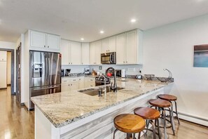 A modern kitchen with white cabinets, stainless steel appliances, a granite countertop, and bar stools. - Lake Daze | Lakefront Powers Lake Home, Sleeps 11 (Genoa City)