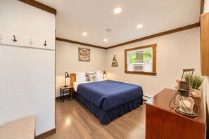 A bedroom with a bed, bedside lamps, a 'NO WAKE ZONE' sign, a window with a view, and a wooden desk with a plant. - Lake Daze | Lakefront Powers Lake Home, Sleeps 11 (Genoa City)