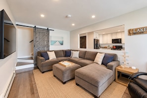 A modern living room with a sectional sofa, a flat-screen TV, a wooden staircase, and a kitchen area. - Lake Daze | Lakefront Powers Lake Home, Sleeps 11 (Genoa City)