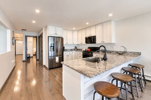 A modern kitchen with white cabinetry, stainless steel appliances, a granite countertop, and bar stools. - Lake Daze | Lakefront Powers Lake Home, Sleeps 11 (Genoa City)