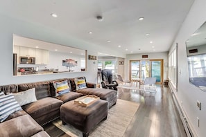 A modern living room with a sofa, ottoman, and a kitchen area in the background. - Lake Daze | Lakefront Powers Lake Home, Sleeps 11 (Genoa City)