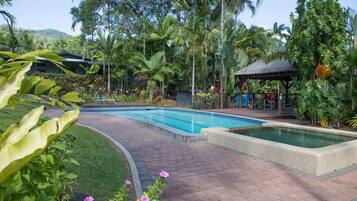 A pool area with a thatched-roof structure, surrounded by lush tropical vegetation and a mountain backdrop.