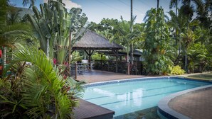 Una piscina rodeada de vegetación tropical, un quiosco con techo de paja y una terraza de madera.