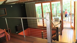 Una escalera de madera conduce a una terraza exterior con barandilla, una pared negra y un ventanal con vistas a un jardín.
