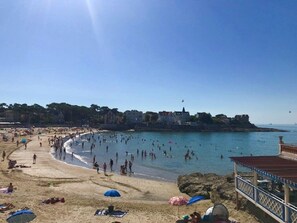 Una playa con gente nadando y tomando el sol, cielo despejado y edificios al fondo.