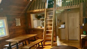 Interior rústico de una cabaña con habitación en entrepiso, muebles de madera, una escalera y zona de cocina.