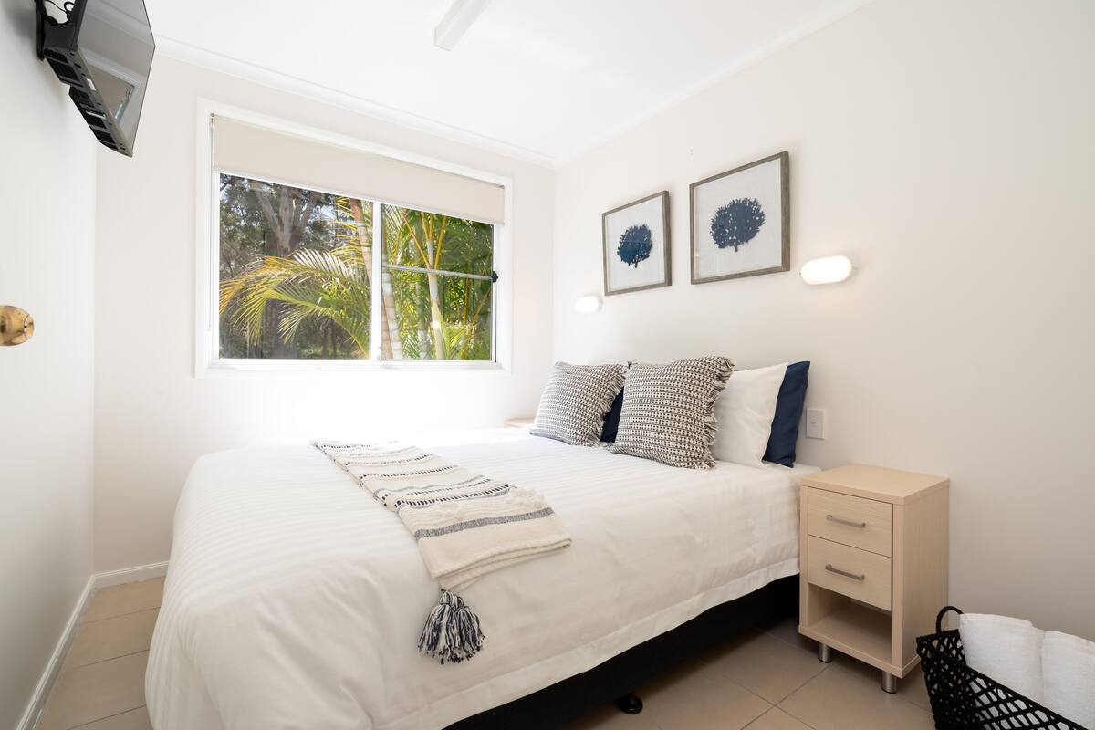 A bedroom with a bed, a nightstand, a window with a view of palm trees, and two wall-mounted artworks.