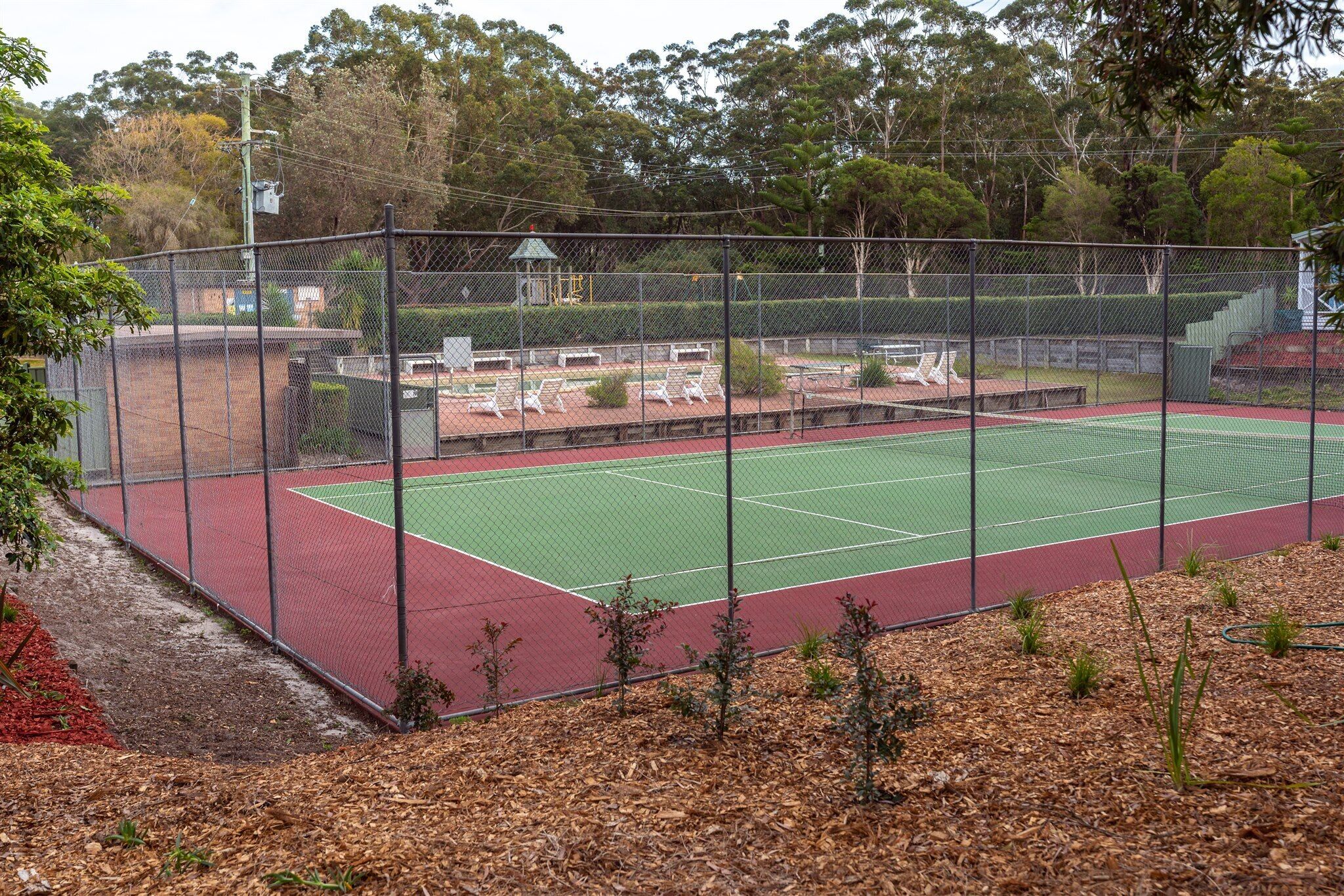 A fenced tennis court with a green playing surface and red borders, surrounded by trees and a small pavilion.