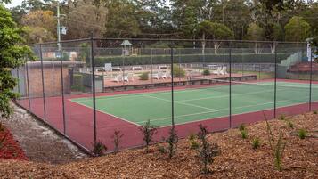 A fenced tennis court with a green playing surface and red borders, surrounded by trees and a small pavilion.