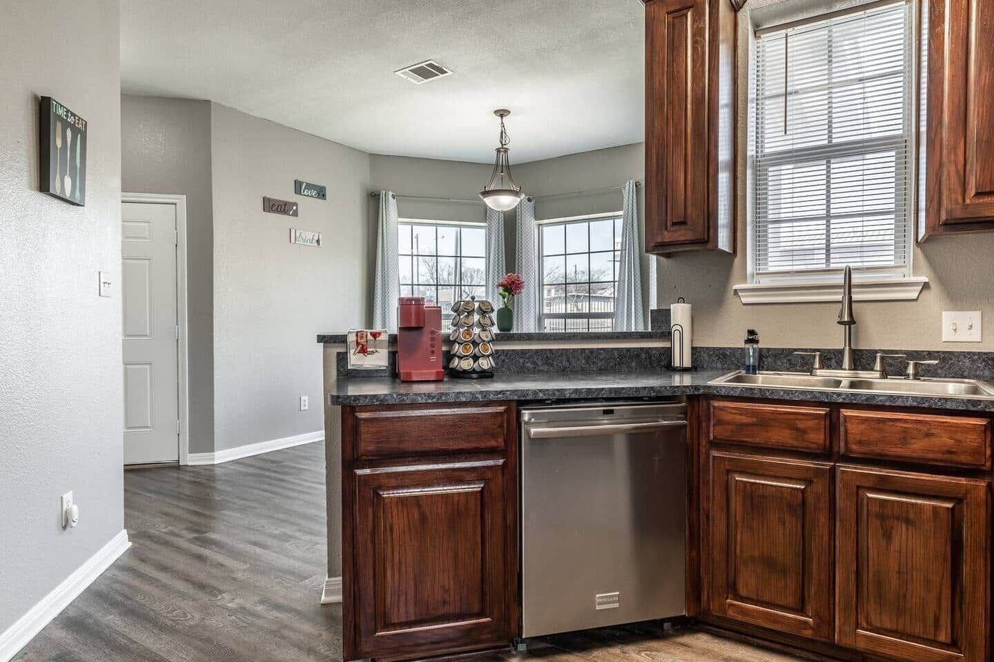 A kitchen with wooden cabinets, a stainless steel dishwasher, a granite countertop, and a window with blinds.