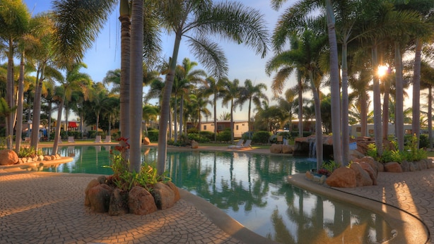 A resort pool area with palm trees, a clear pool, and a paved walkway. - Tasman Holiday Parks - Rollingstone (Rollingstone)