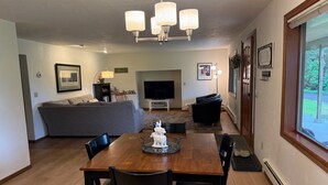 A dining area with a wooden table and chairs, a grey sofa, a TV, and a window with a view of greenery. - A Cozy Home Away From Home in Alaska! (Juneau)