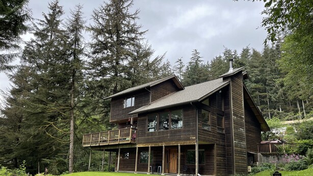 A two-story wooden cabin with a balcony, surrounded by dense forest. - A Cozy Home Away From Home in Alaska! (Juneau)