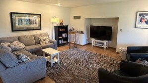 A living room with a grey sofa, a small white table, a black armchair, a TV on a stand, a bookshelf, and a framed picture on the wall. - A Cozy Home Away From Home in Alaska! (Juneau)