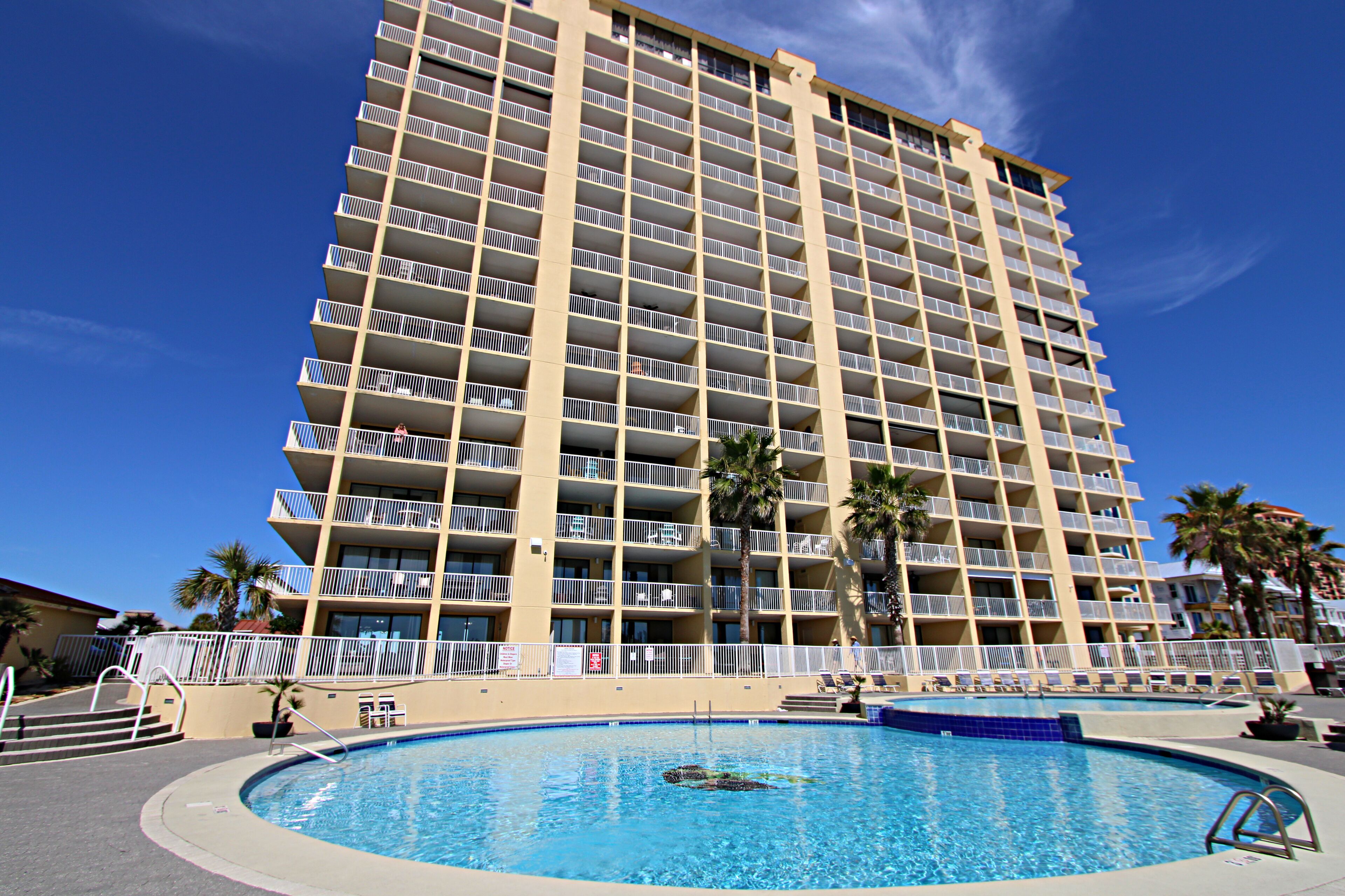 A high-rise hotel with a swimming pool, multiple balconies, and palm trees.
