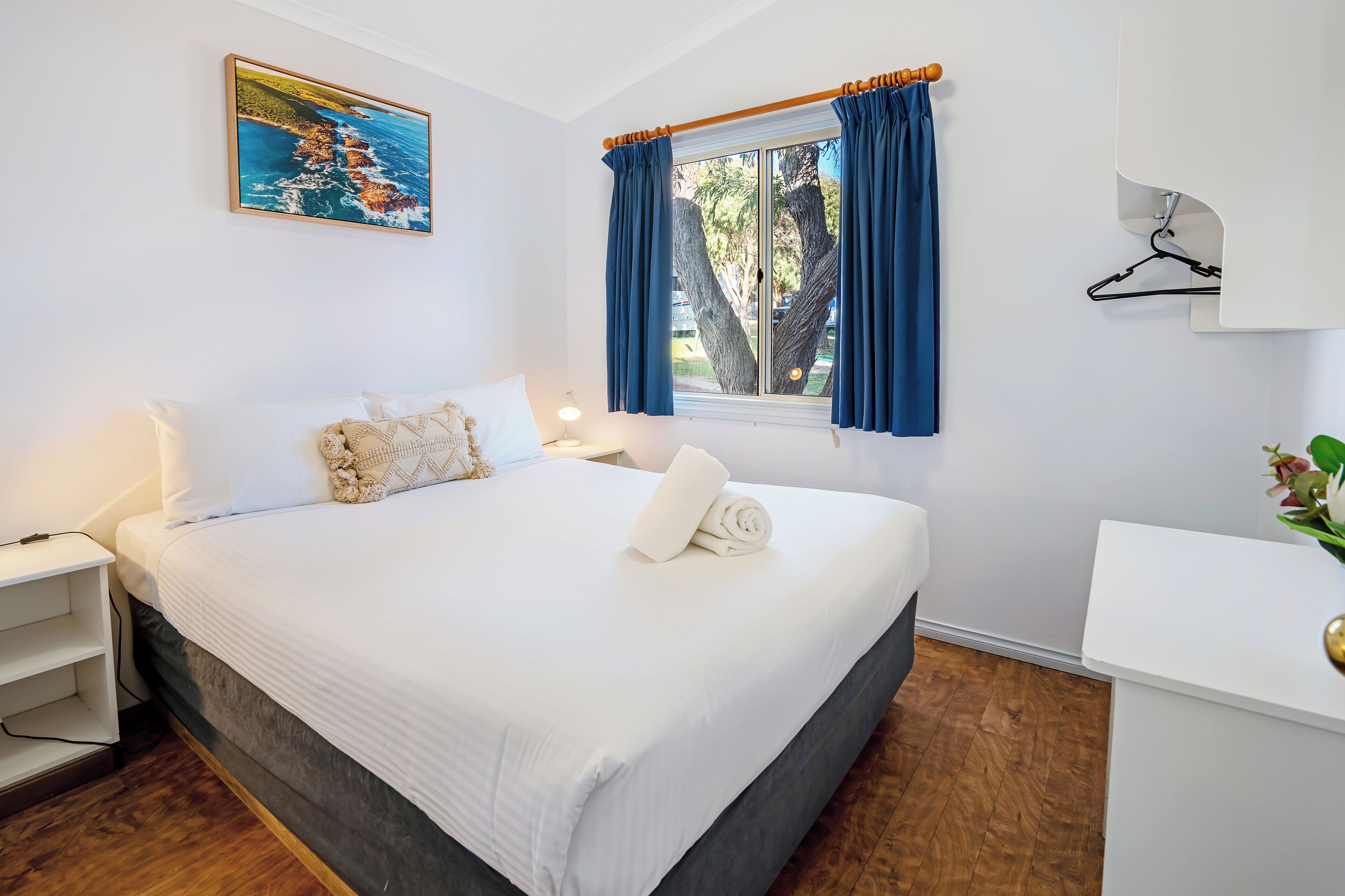 A neatly made bed with white linens, a bedside table with a lamp, a window with blue curtains, and a painting of a coastal scene on the wall.