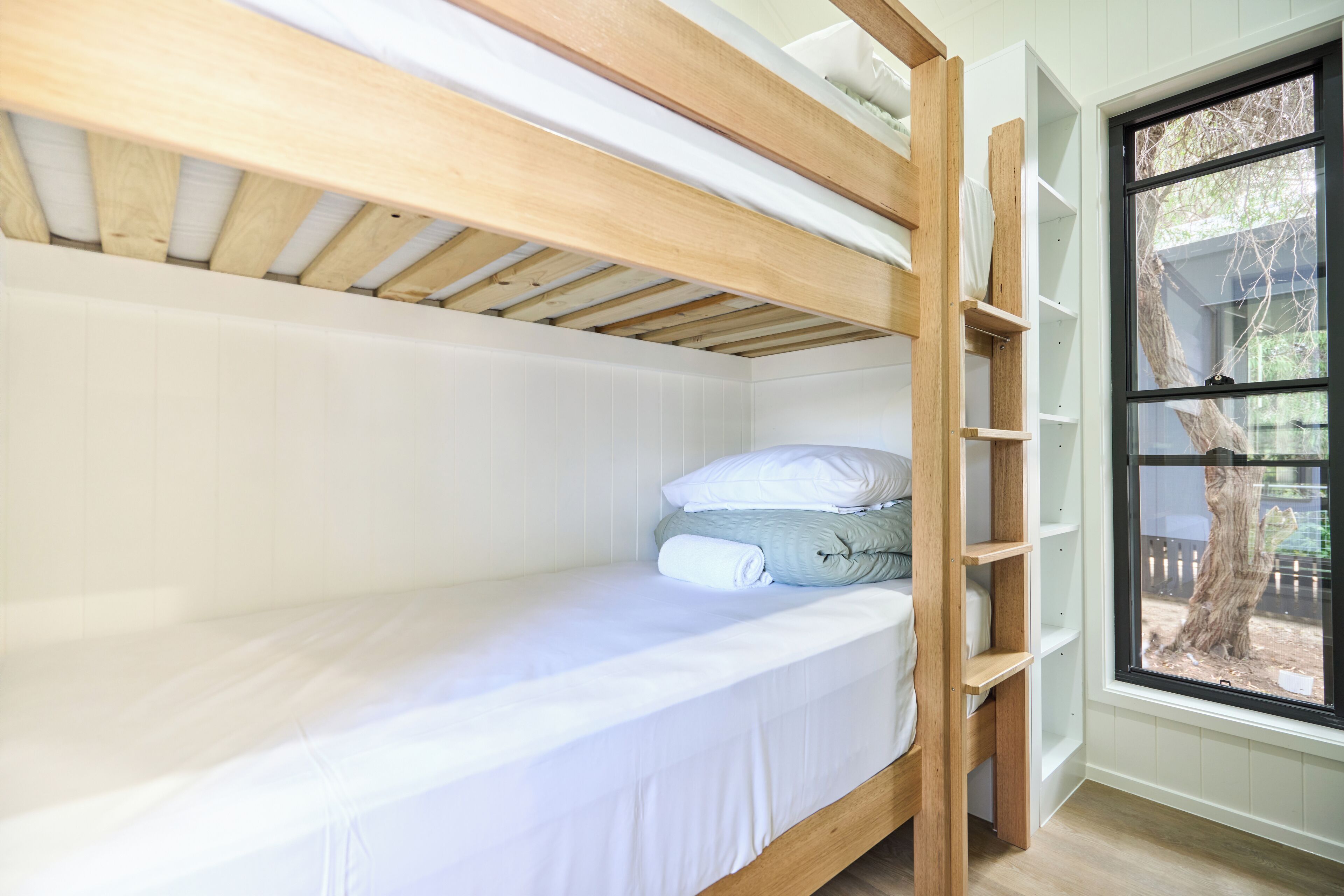 A bunk bed with a wooden frame and white bedding. There is a window with a view of a tree and a fence.