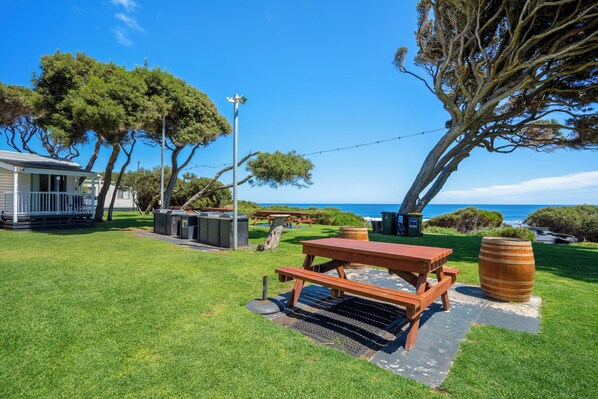 A picnic area with a wooden table and benches, a barbecue grill, and a large barrel, surrounded by trees and a view of the ocean. - Tasman Holiday Parks - Yallingup Beach (Yallingup)