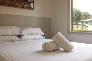 Una cama bien hecha con sábanas y almohadas blancas, una toalla blanca enrollada y una ventana con vistas a árboles y un edificio.