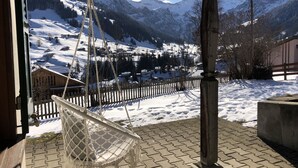 Ein Hängestuhl auf einer Ziegelterrasse mit Blick auf eine verschneite Berglandschaft, mit Holzzaun und Gebäuden im Tal.