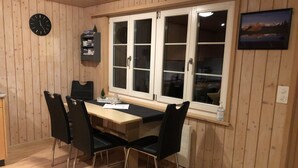 Ein Zimmer mit Holzwandverkleidung, einem Esstisch, vier Stühlen, einer Uhr, einem Fenster mit weißen Rahmen und einem Bild in einem Rahmen an der Wand.
