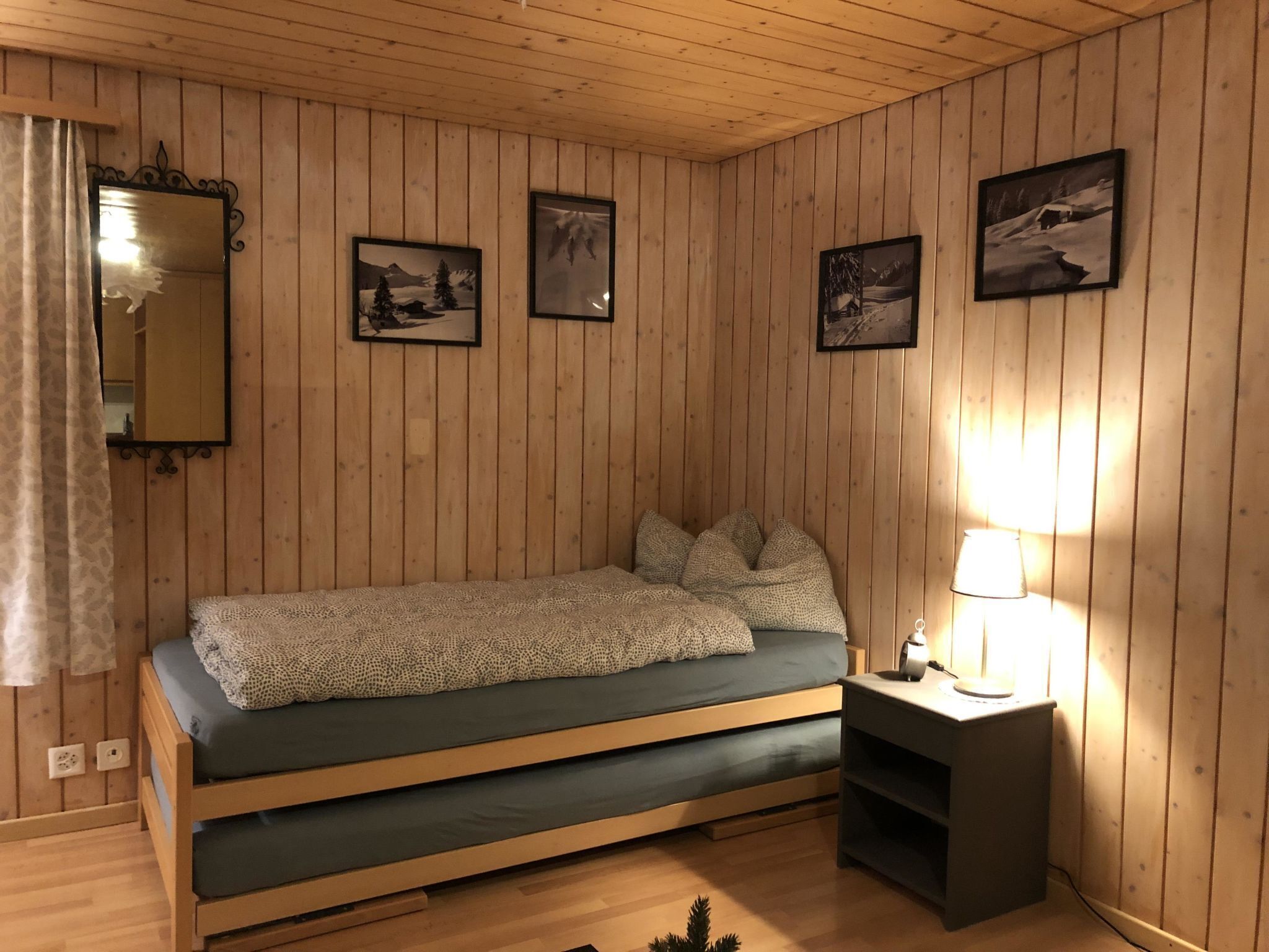 A cozy room with wooden walls, a bed with a patterned blanket, a bedside lamp, and framed pictures on the wall.
