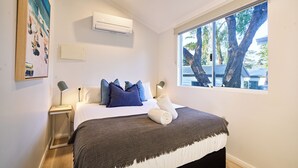 A bedroom with a bed, bedside tables, an air conditioner, a window with a view of trees, and a painting on the wall. - Tasman Holiday Parks - Yallingup Caves (Yallingup)
