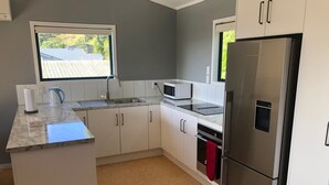 A modern kitchen with white cabinets, a stainless steel refrigerator, a microwave, and a built-in oven. - The Kakariki Cottage - Experience all that Aotea has to offer (Aotea, Kawhia)