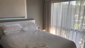 A neatly made bed with white linens and two pillows, a window with a view of greenery, and a balcony with sheer curtains. - The Kakariki Cottage - Experience all that Aotea has to offer (Aotea, Kawhia)