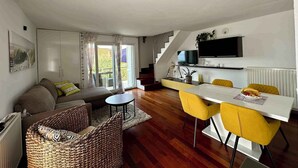 Een moderne woonkamer met een bank, een eettafel met gele stoelen, een trap en een aan de muur bevestigde televisie.