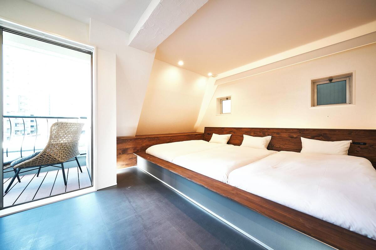 Una habitación de hotel moderna con una cama grande, cabecera de madera, un ventanal pequeño y un balcón con una silla de mimbre.