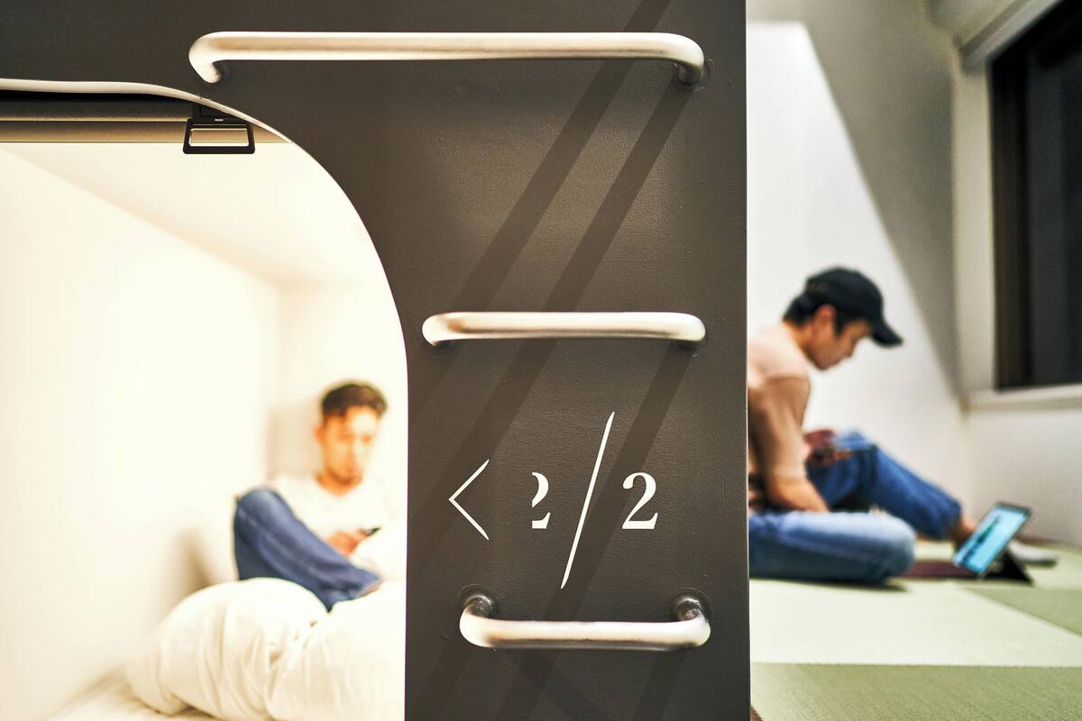 Una habitación con cama, ventana y una persona usando una computadora portátil.