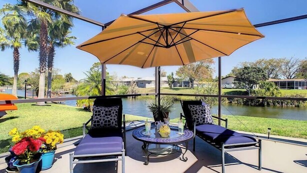 A covered patio with two chairs and a table, overlooking a body of water and a grassy area with palm trees.