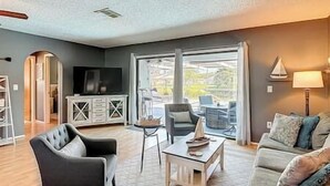A living room with a sofa, armchair, coffee table, TV, and a view through large windows.