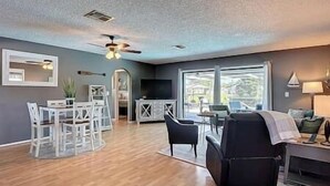 A spacious living room with a wooden floor, a ceiling fan, a dining area with white chairs, and a large window offering a view of the outdoors.
