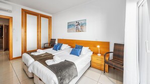 A hotel room with a large bed, a wooden headboard, a painting on the wall, a wicker chair, and a nightstand with a lamp.