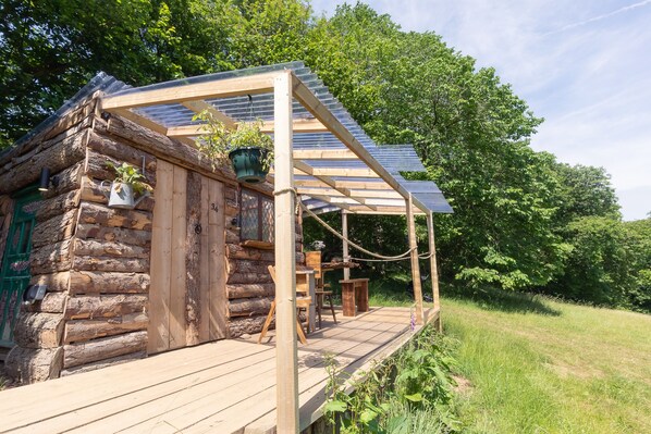 A rustic log cabin with a wooden deck, a pergola with a sloped roof, and a hanging planter. - Wood Cabin & 4 Large Bell Tents - Hapus Hideaway (Abergele)