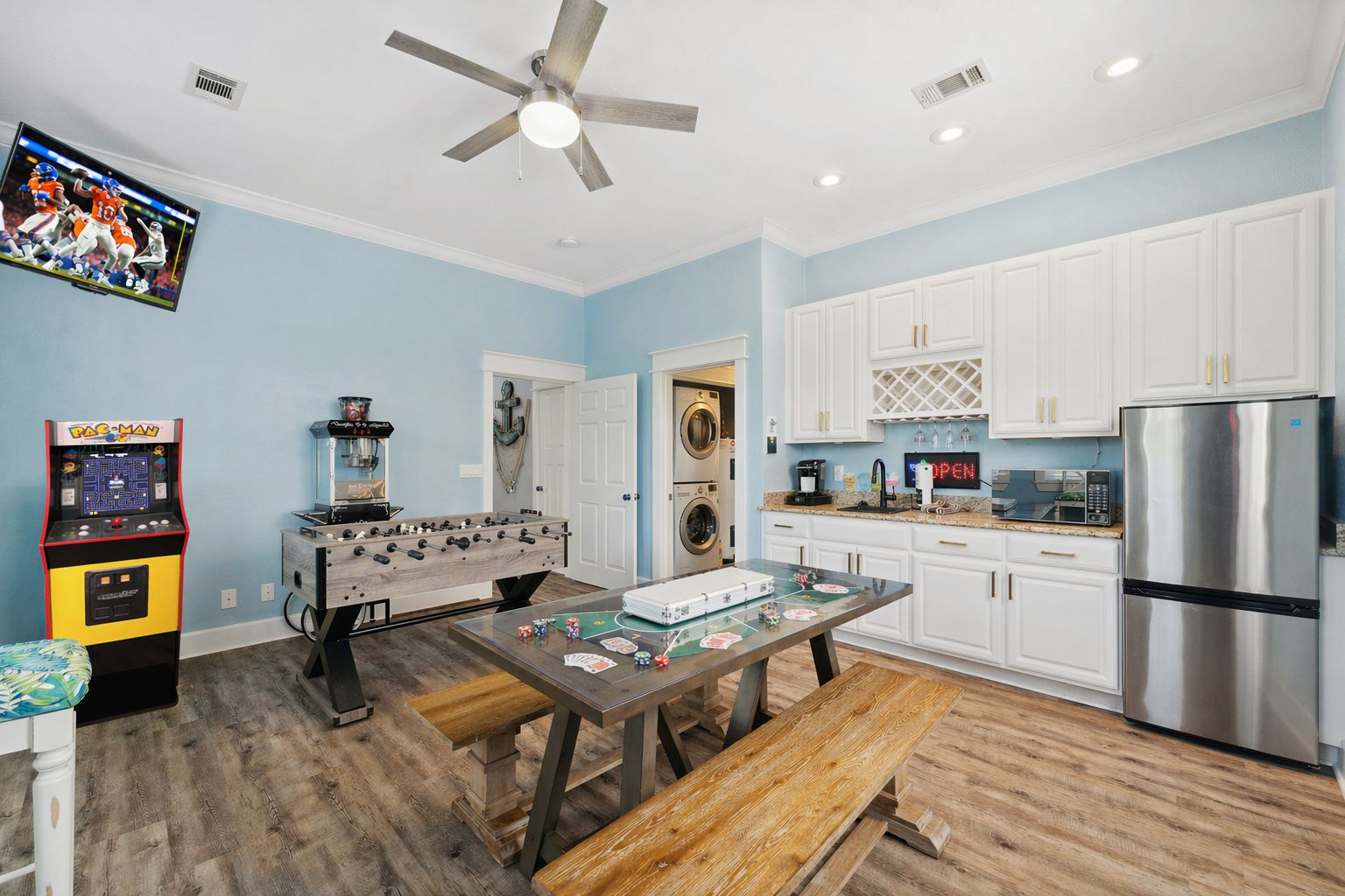 A kitchen with white cabinets, a stainless steel refrigerator, a foosball table, and a Pac-Man arcade game.