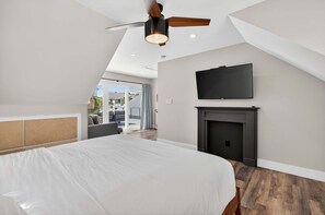 A bedroom with a large bed, a ceiling fan, a fireplace, and a TV mounted on the wall.