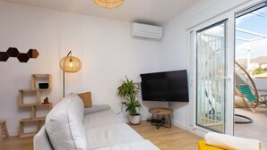 Ein modernes Wohnzimmer mit einer Couch, einem gelben Sessel, einem Fernseher und einem großen Fenster mit Blick ins Freie.