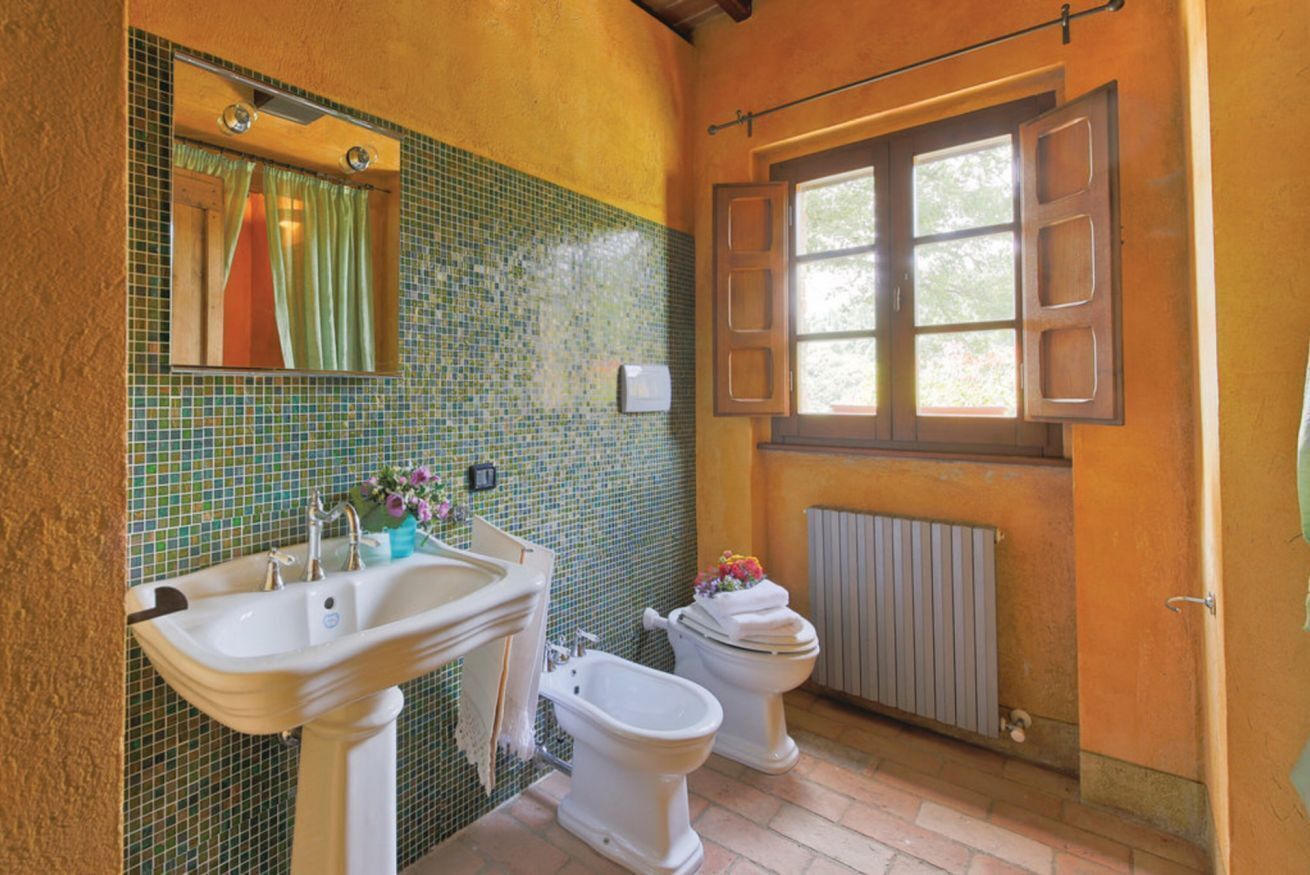 Une salle de bain avec un lavabo, deux toilettes, un miroir et une fenêtre dont les volets sont ouverts.
