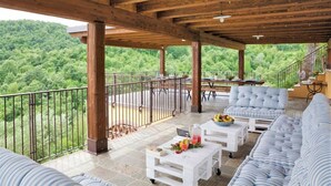 A covered outdoor seating area with a view of a forested hillside, wooden beams, and a white table with a fruit bowl.