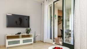 A living room with a flat-screen TV, a white console table with decorative items, a glass door leading to a balcony, and a small table with a bowl of fruit and two glasses. - Lovely apartment for 4 guests with WIFI, A/C, TV and balcony (Tossa De Mar)