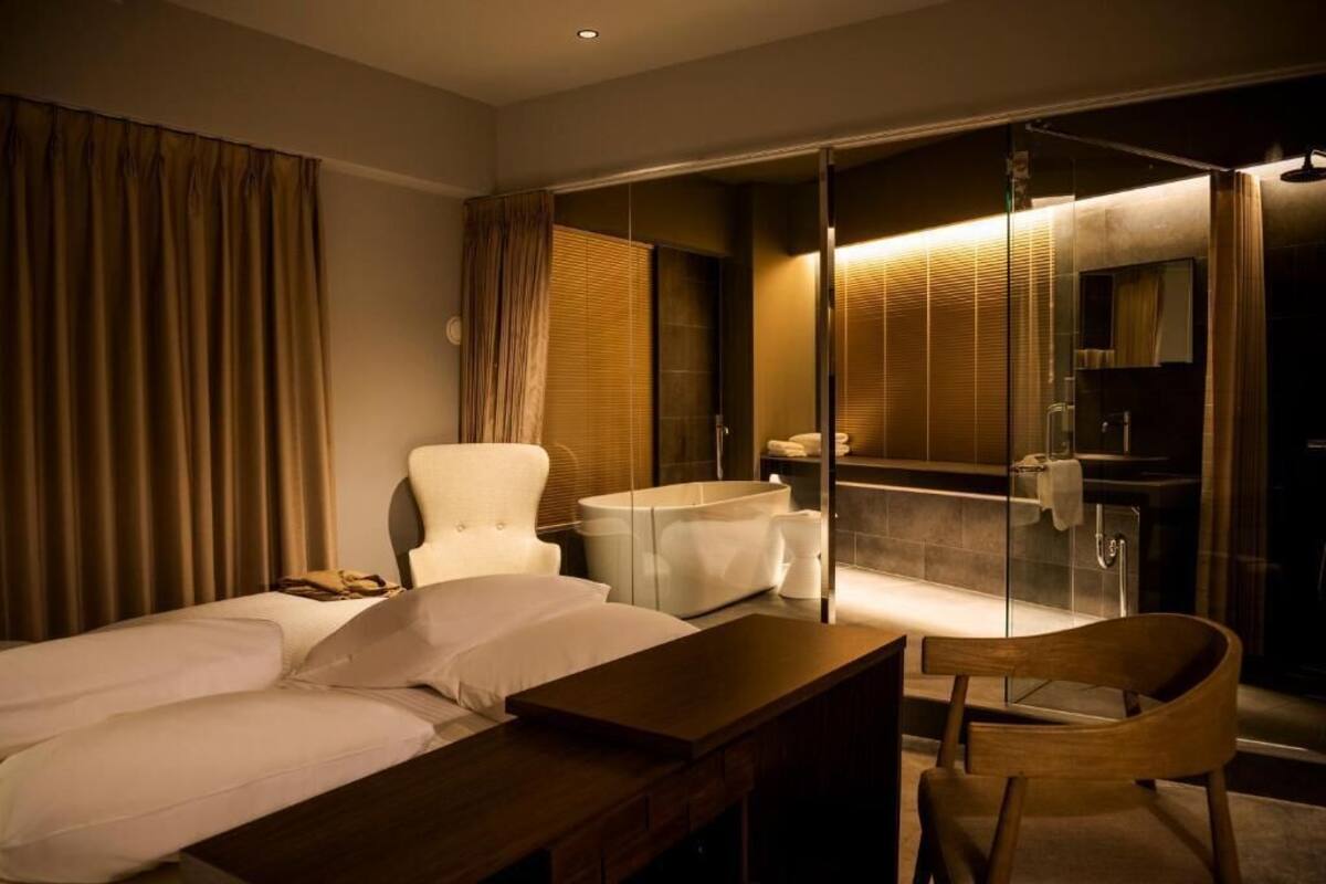 A hotel room with a bed, a wooden desk, a chair, a bathroom with a glass shower enclosure, and a large mirror.